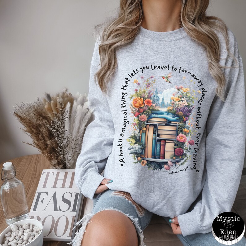 K&ouml;nnte beinhalten: Ein graues Sweatshirt mit einem grafischen Druck eines Stapels B&uuml;cher, umgeben von Blumen und einem Zitat, das lautet: "Ein Buch ist eine magische Sache, die es dir erm&ouml;glicht, an ferne Orte zu reisen, ohne jemals wegzugehen" von Katrina Mayer.