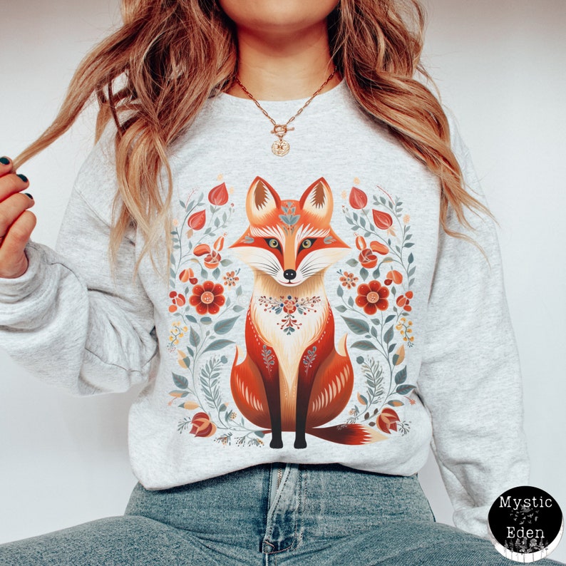 Op de afbeelding: Een grijs sweatshirt met een kleurrijke illustratie van een vos omringd door bloemen. De vos is oranje, rood en wit met een bloemenkraag. De bloemen zijn rood, oranje, geel en groen.