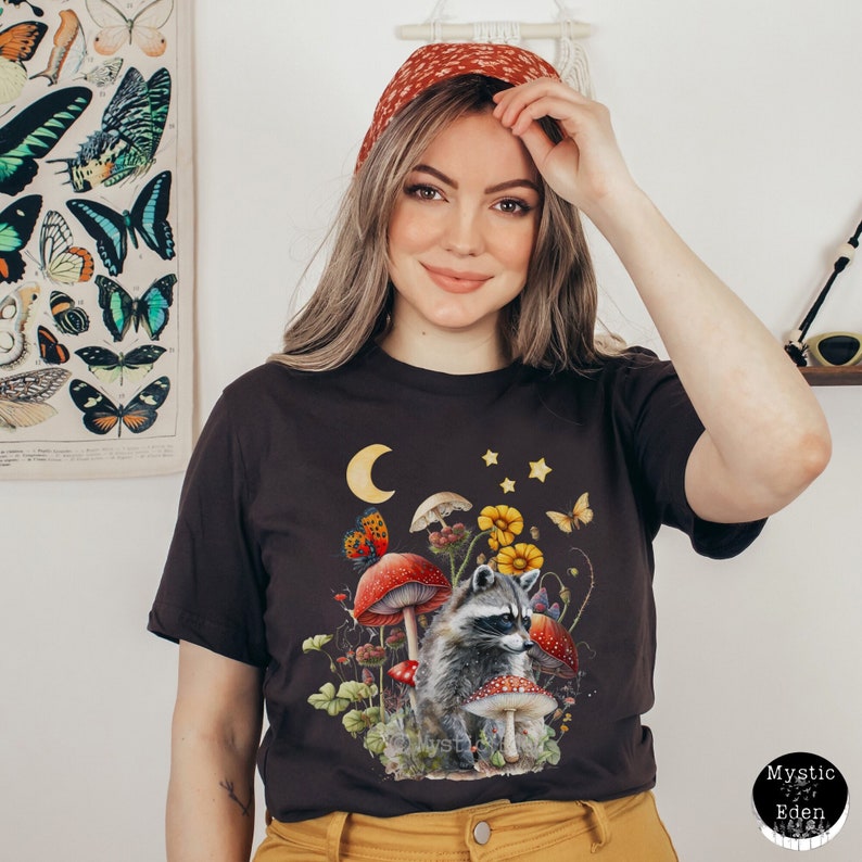 May include: A dark brown t-shirt with a graphic of a raccoon sitting amongst mushrooms, flowers, and butterflies. The moon and stars are in the background.