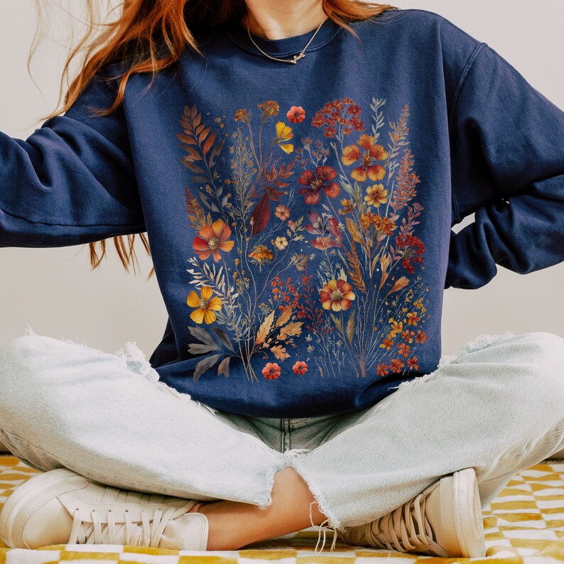 May include: A navy blue sweatshirt featuring a watercolor floral design in shades of orange, yellow, and red. The botanical illustration includes various flowers and foliage. The sweatshirt is a casual garment, suitable for everyday wear.