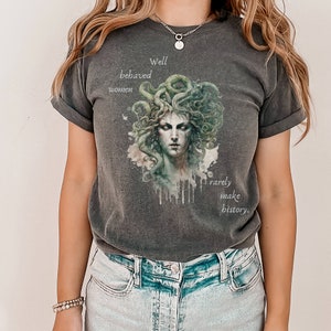May include: Dark grey t-shirt featuring a Medusa illustration with green and white accents. The text reads "Well behaved women rarely make history." The shirt has a classic crew neck and short sleeves.