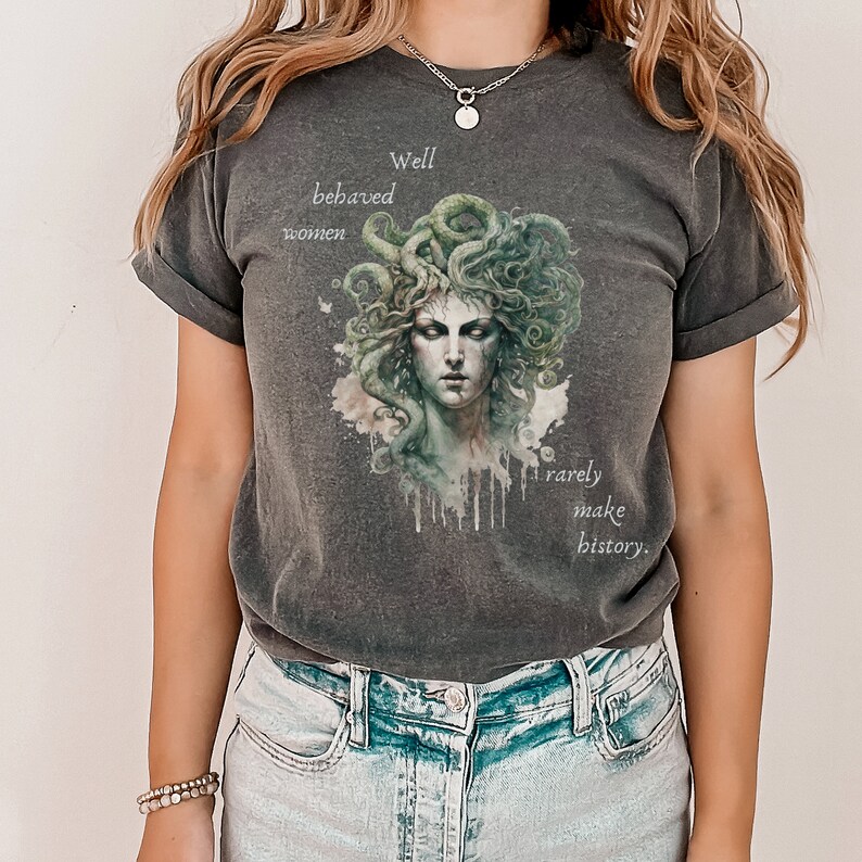 May include: Dark grey t-shirt featuring a Medusa illustration with green and white accents. The text reads "Well behaved women rarely make history." The shirt has a classic crew neck and short sleeves.