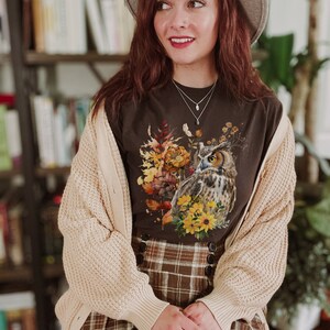 May include: A woman wearing a brown cardigan over a brown t-shirt with a graphic print of an owl and fall foliage. The t-shirt has a brown and white plaid skirt.