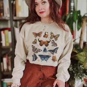 May include: A beige sweatshirt with a print of various butterflies in shades of brown, orange, black, and blue.