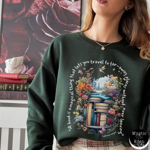 K&ouml;nnte beinhalten: Ein dunkelgr&uuml;ner Pullover mit einem wei&szlig;en Grafikdruck eines B&uuml;cherstapels, umgeben von Blumen und einem Kolibri. Der Text auf der Grafik lautet "A book is a magical thing that lets you travel to far-away places without ever leaving". Der Text "katrina mayer" ist ebenfalls in der Grafik enthalten.