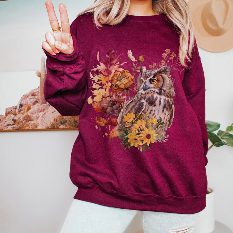 May include: A maroon sweatshirt with a graphic print of an owl surrounded by fall foliage and flowers. The owl is brown and gray with yellow eyes. The flowers are yellow and orange.