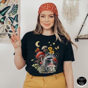 May include: Black t-shirt with a graphic of a raccoon in a forest setting with mushrooms, flowers, and a crescent moon. The raccoon is wearing a red bandana.