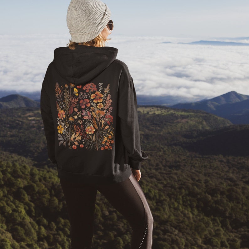May include: A black hoodie with a floral print on the back. The hoodie has a drawstring hood and a kangaroo pocket. The person wearing the hoodie is standing on a mountaintop with a view of clouds and mountains in the distance.