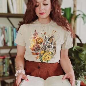 May include: A beige t-shirt with a graphic print of an owl and fall foliage. The owl is brown and white with yellow eyes. The foliage is in shades of orange, yellow, and brown.