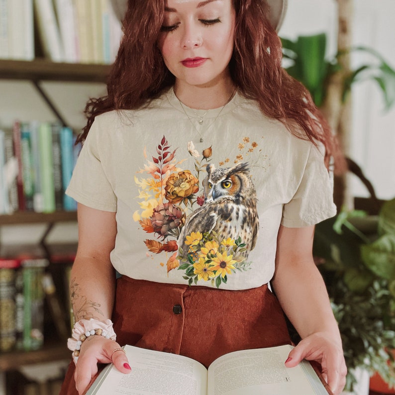 May include: A beige t-shirt with a graphic print of an owl and fall foliage. The owl is brown and white with yellow eyes. The foliage is in shades of orange, yellow, and brown.