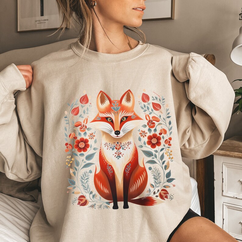 Op de afbeelding: Een beige sweatshirt met een grafische print van een vos omringd door bloemen. De vos is oranje en wit met een bloemmotief om zijn nek.