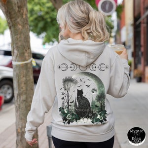 May include: A light gray hooded sweatshirt with a black cat silhouette in a moonlit forest scene. The design features a crescent moon, stars, and floral elements. The text "The Mystical" is printed on the cat's back.