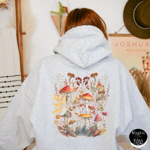 Op de afbeelding: Grijs hoodie met een kleurrijke paddenstoel illustratie op de rug. De illustratie toont een verscheidenheid aan paddenstoelen in tinten rood, oranje, geel en bruin, omringd door groene bladeren en bloemen.
