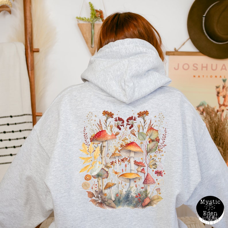 Op de afbeelding: Grijs hoodie met een kleurrijke paddenstoel illustratie op de rug. De illustratie toont een verscheidenheid aan paddenstoelen in tinten rood, oranje, geel en bruin, omringd door groene bladeren en bloemen.
