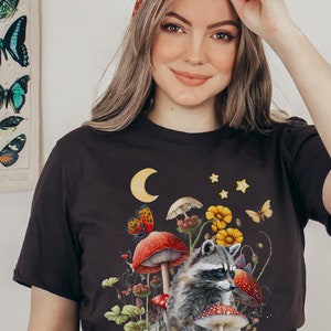 May include: A black t-shirt with a graphic print of a raccoon sitting in a field of mushrooms and flowers. The graphic also includes a crescent moon and stars.