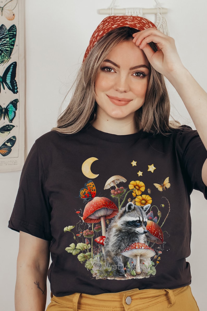 May include: A black t-shirt with a graphic print of a raccoon sitting in a field of mushrooms and flowers. The graphic also includes a crescent moon and stars.