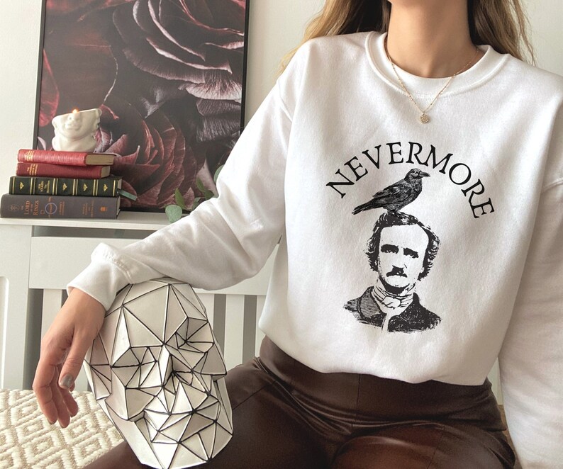 May include: White sweatshirt with a black and white illustration of Edgar Allan Poe with a raven perched on his head. The text "NEVERMORE" is printed in a black arc around the illustration.