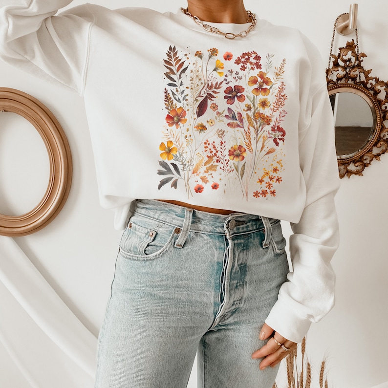 May include: White cropped sweatshirt with a floral print featuring orange, yellow, and red flowers and leaves. The sweatshirt is worn with light blue jeans.