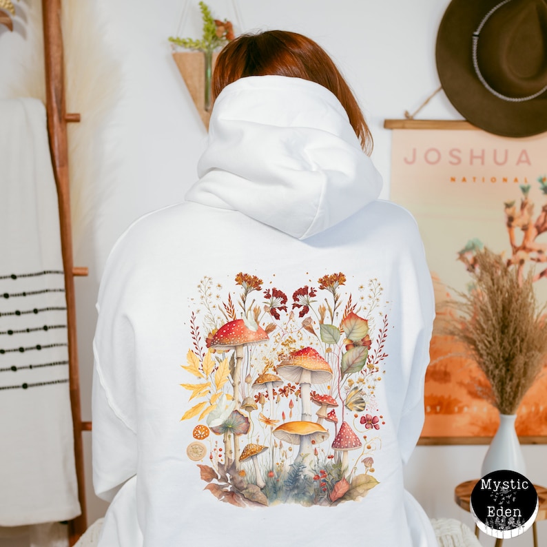 Op de afbeelding: Witte hoodie met een kleurrijke paddenstoelenillustratie op de rug. De illustratie toont rode, oranje en gele paddenstoelen met groene bladeren en bloemen.