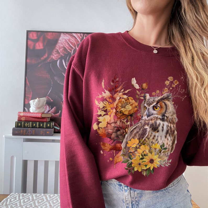 May include: A maroon sweatshirt with a graphic print of an owl surrounded by fall foliage and flowers. The owl has brown and white feathers and yellow eyes.