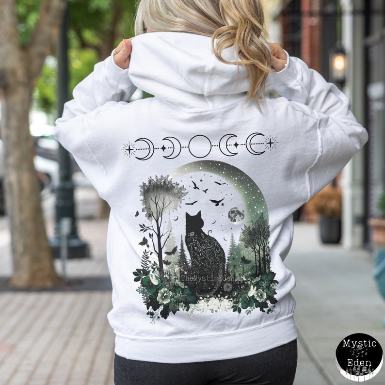 May include: White hooded sweatshirt with a black cat silhouette sitting in a moonlit forest with green foliage and flowers. The moon is surrounded by a crescent moon phase design and a star. The text "The Mystic Eden" is visible at the bottom of the design.