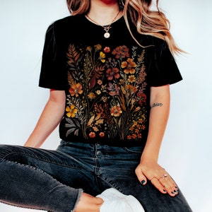 Puede incluir: Una camiseta negra con un estampado floral que presenta flores secas en tonos naranja, amarillo y marr&oacute;n. Las flores est&aacute;n dispuestas en un dise&ntilde;o de ramo.
