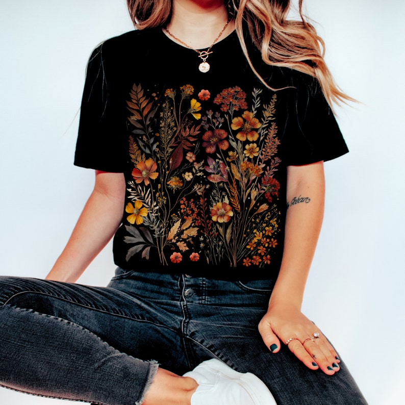 Puede incluir: Una camiseta negra con un estampado floral que presenta flores secas en tonos naranja, amarillo y marr&oacute;n. Las flores est&aacute;n dispuestas en un dise&ntilde;o de ramo.