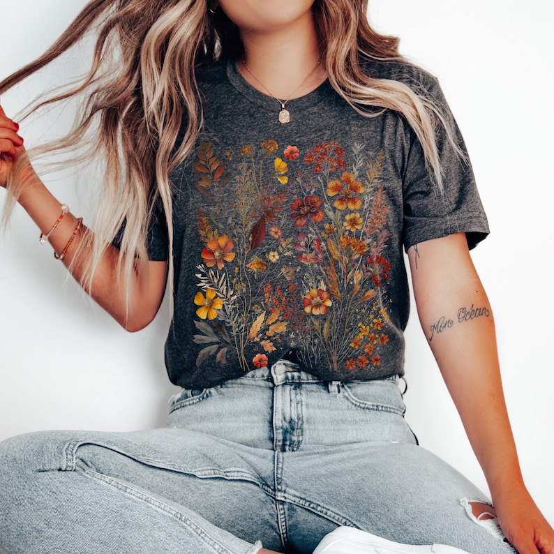 Puede incluir: Una camiseta gris antracita con un estampado floral que presenta flores secas en tonos naranja, amarillo y rojo. Las flores est&aacute;n dispuestas en un dise&ntilde;o similar a un ramo.