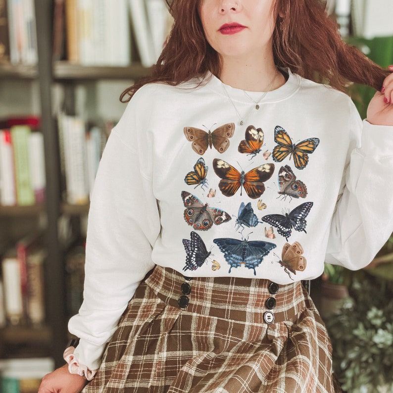 May include: White sweatshirt with a colorful butterfly print. The butterflies are arranged in a random pattern on the front of the shirt. The shirt is worn by a person with long brown hair and is paired with a brown and white plaid skirt.