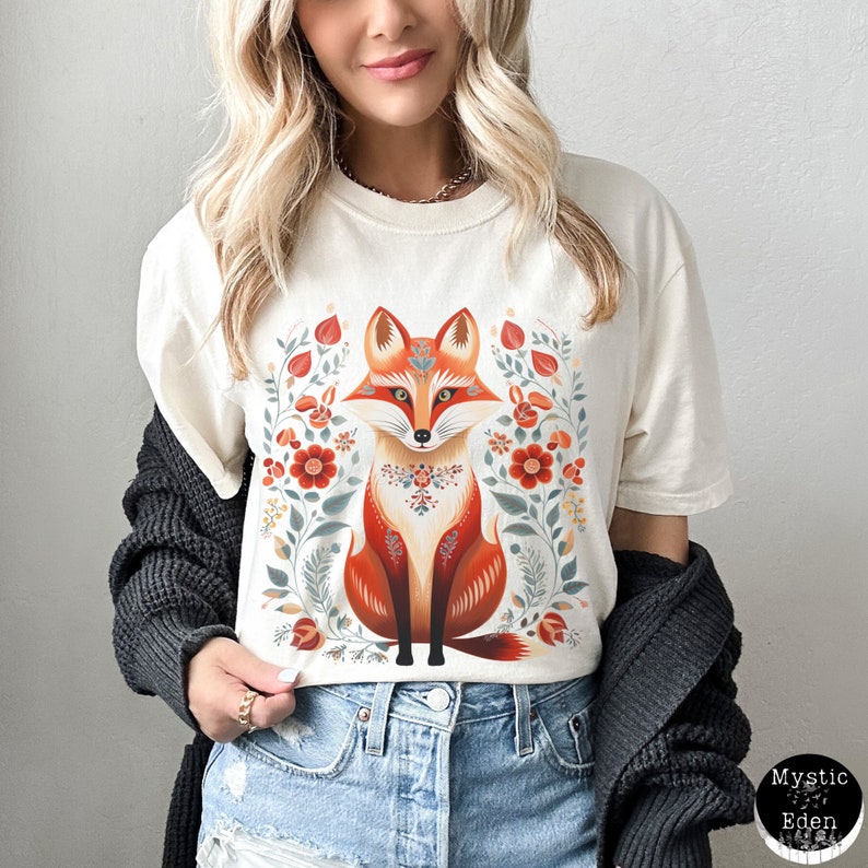 May include: Cream-coloured t-shirt with a stylized fox illustration. The fox is in shades of orange, red, and brown, with floral and leaf accents. The design includes a floral border and decorative patterns. The shirt has a relaxed fit.