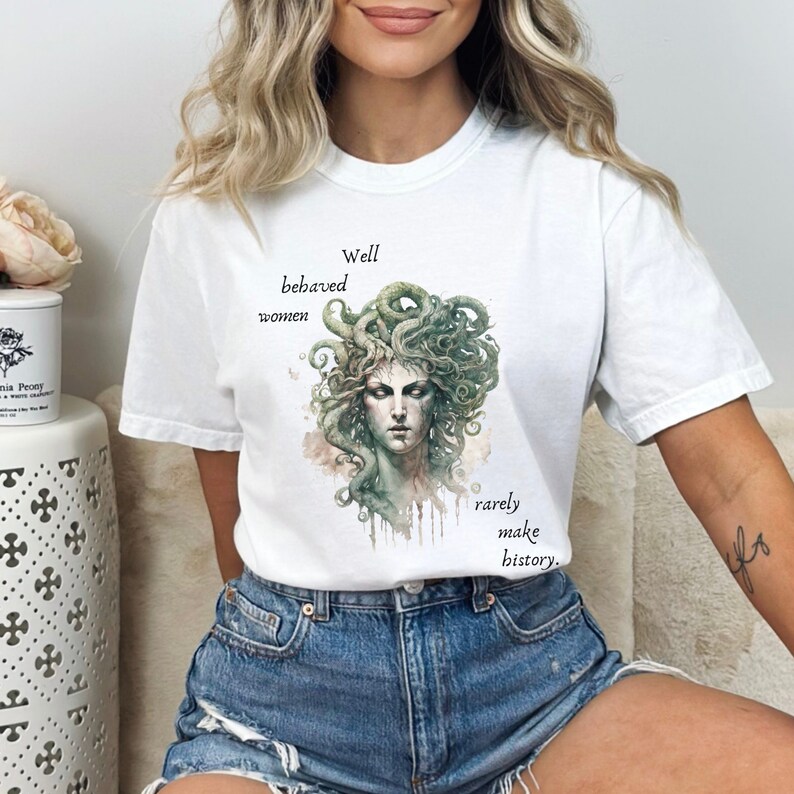 May include: White t-shirt featuring a watercolor illustration of Medusa with green snakes for hair. The text "Well behaved women rarely make history" is printed on the shirt in black.