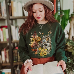 May include: A dark green sweatshirt with a floral print featuring an owl and autumn leaves. The sweatshirt has a crew neck and long sleeves.