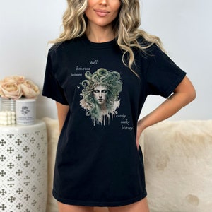 May include: Black t-shirt featuring a watercolor illustration of Medusa with green snakes for hair. The text reads "Well behaved women rarely make history." The shirt is a comfortable, casual style.