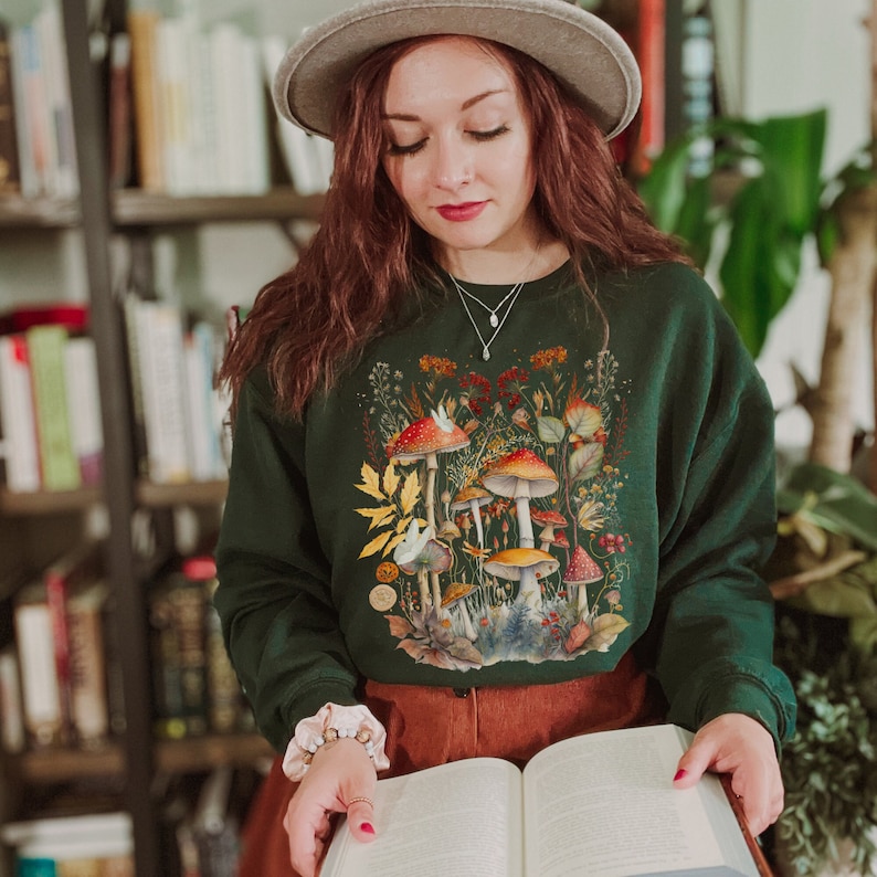 Pode incluir: Uma mulher vestindo uma camisa de frio verde com uma estampa de cogumelos e o texto "Shroom Queens". Ela est&aacute; segurando um livro em suas m&atilde;os.