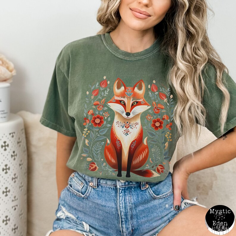 May include: An olive green t-shirt with a fox illustration in the centre, featuring red and orange fur and floral details. The shirt is worn with denim shorts. The Mystic Eden logo is visible on the bottom right of the image.
