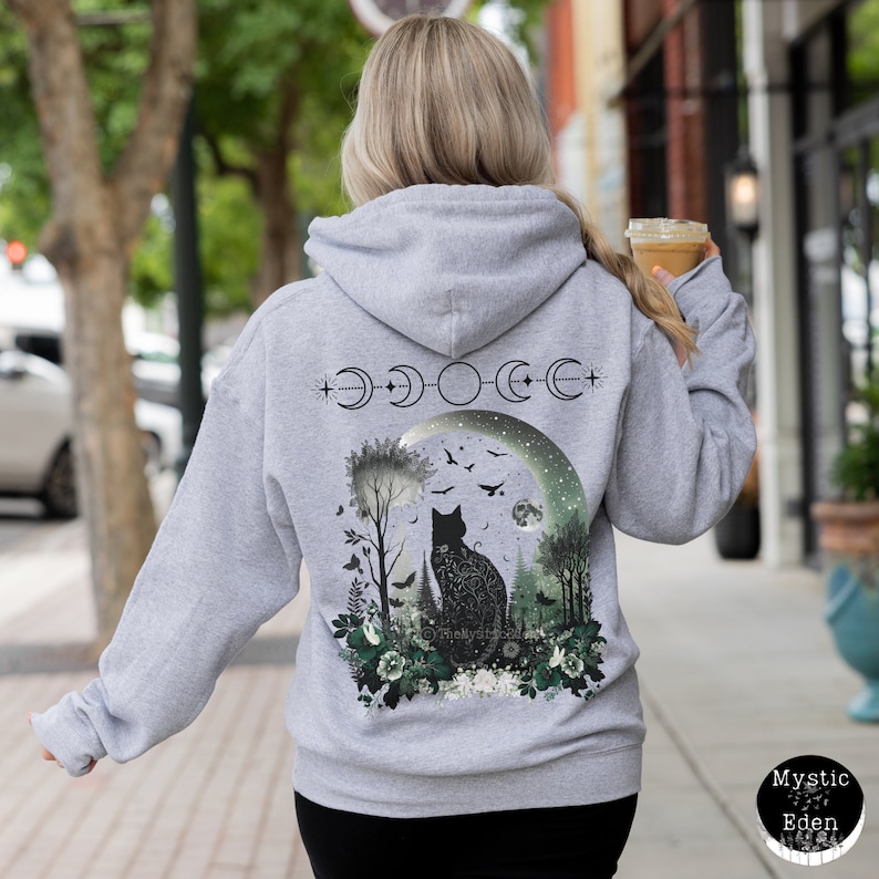 May include: Gray hooded sweatshirt with a black cat silhouette in a moonlit forest scene. The cat is surrounded by flowers and trees. The moon is surrounded by a crescent moon phase design. The text "Mystic Eden" is visible in the lower right corner.