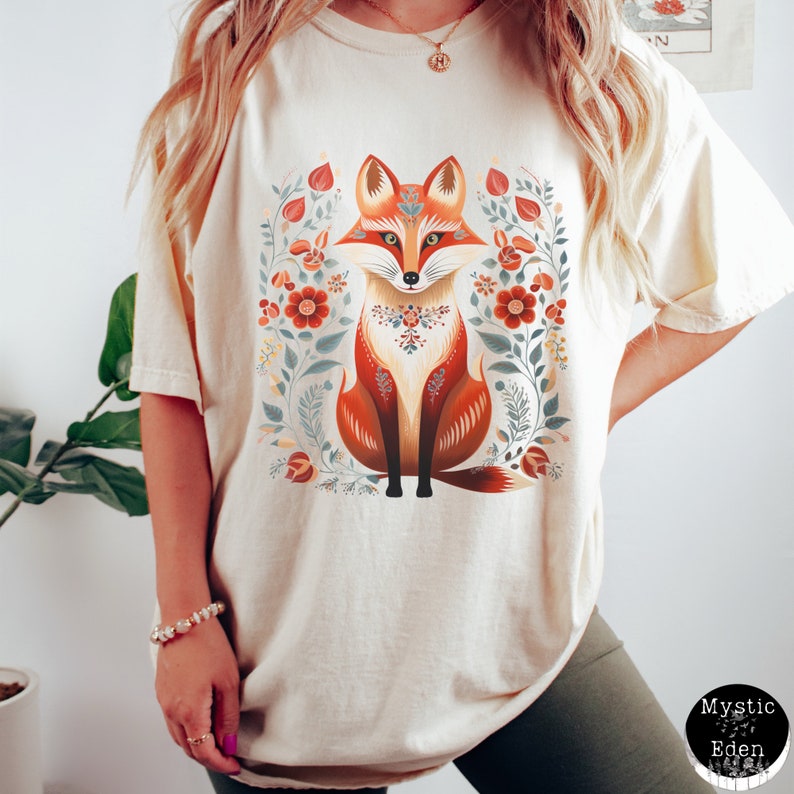 May include: A cream-coloured t-shirt with a detailed illustration of a fox surrounded by floral elements. The fox is in shades of orange, brown, and white, with intricate patterns. The floral design includes red and blue flowers. The shirt has a relaxed fit.