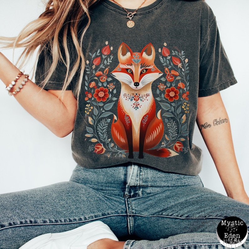 May include: Dark grey t-shirt with a fox illustration. The fox is in orange, red, and white, surrounded by red, yellow, and green floral designs. The shirt has a vintage, washed appearance.
