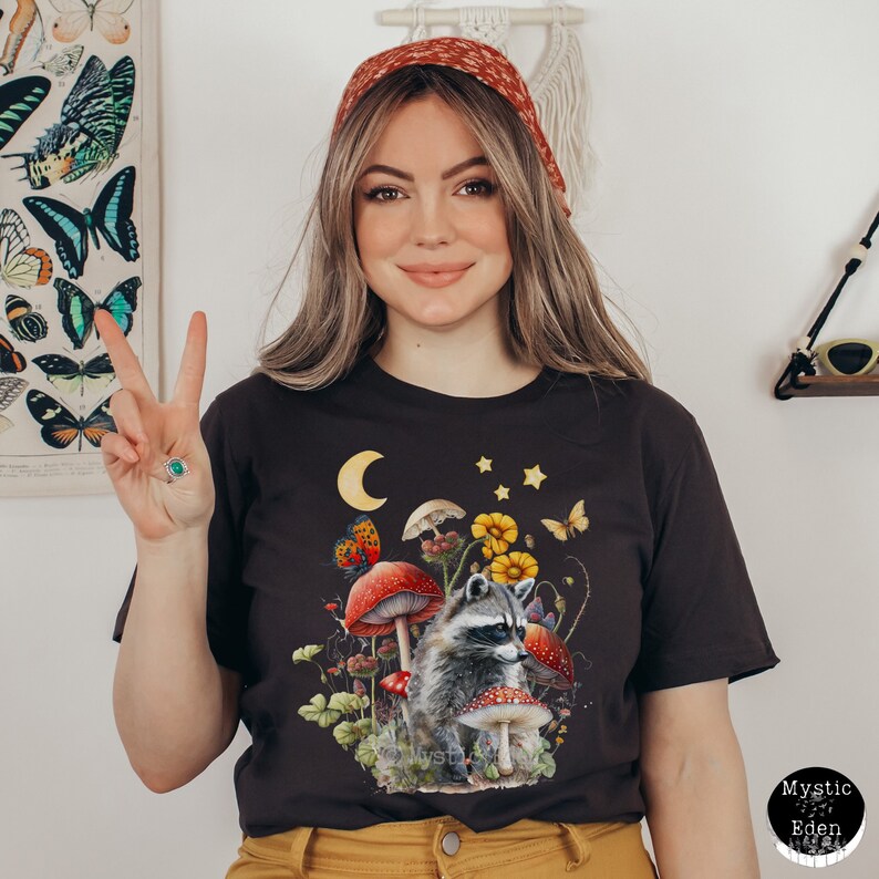 May include: A dark grey t-shirt with a graphic of a raccoon sitting amongst mushrooms, butterflies, and flowers. The graphic also includes a crescent moon and stars. The text "Mystic Eden" is visible in the bottom right corner of the image.