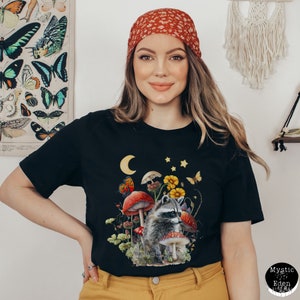 May include: Black t-shirt with a graphic of a raccoon in a mushroom forest. The raccoon is surrounded by mushrooms, flowers, and butterflies. The moon and stars are visible in the sky.