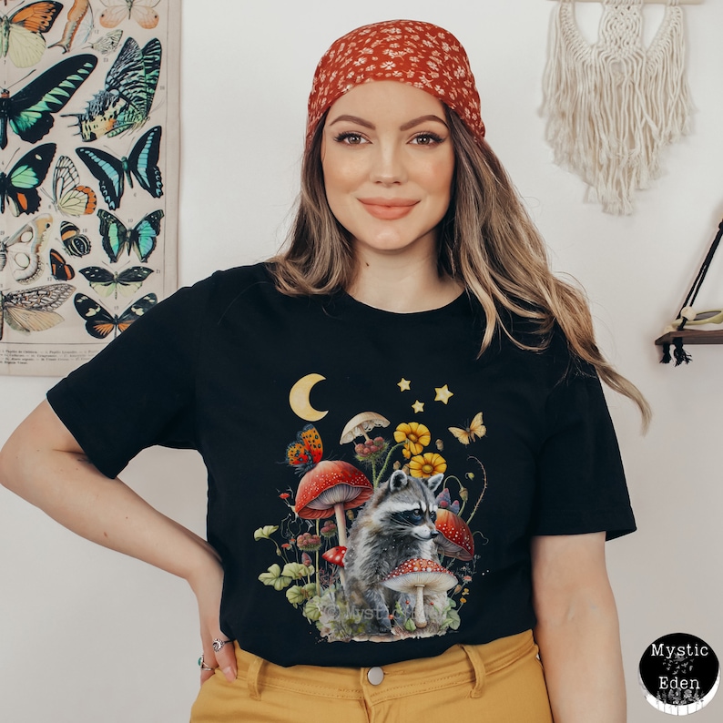 May include: Black t-shirt with a graphic of a raccoon in a mushroom forest. The raccoon is surrounded by mushrooms, flowers, and butterflies. The moon and stars are visible in the sky.