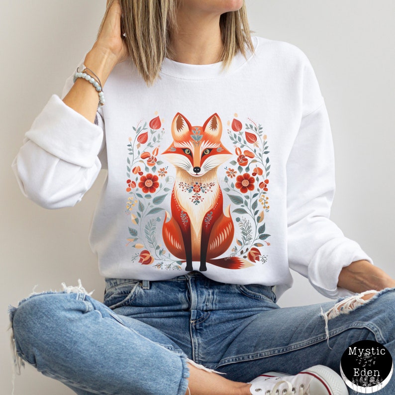 Op de afbeelding: Witte trui met een rode vos-illustratie omringd door bloemmotieven. De vos zit met zijn staart om zijn poten gewikkeld. De bloemen zijn in tinten rood, roze en wit.
