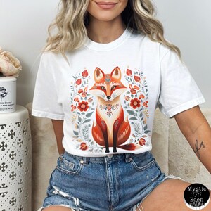 May include: White t-shirt with a detailed fox design. The fox is mainly orange with decorative patterns, set within a floral wreath of red, orange, and green. The tee has short sleeves and a loose fit, suitable for casual wear.