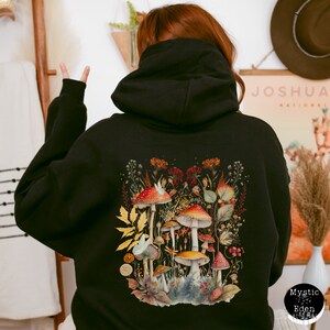 Op de afbeelding: Zwarte hoodie met een aquarel-illustratie van paddenstoelen en bladeren op de rug. De illustratie heeft rode, oranje, gele en groene kleuren. De tekst "Mystic Eden" is gedrukt in de rechterbenedenhoek van de hoodie.