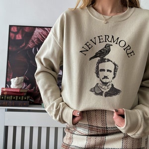 May include: A beige sweatshirt with a black graphic print of Edgar Allan Poe with a raven perched on his head. The text "NEVERMORE" is printed above the graphic.