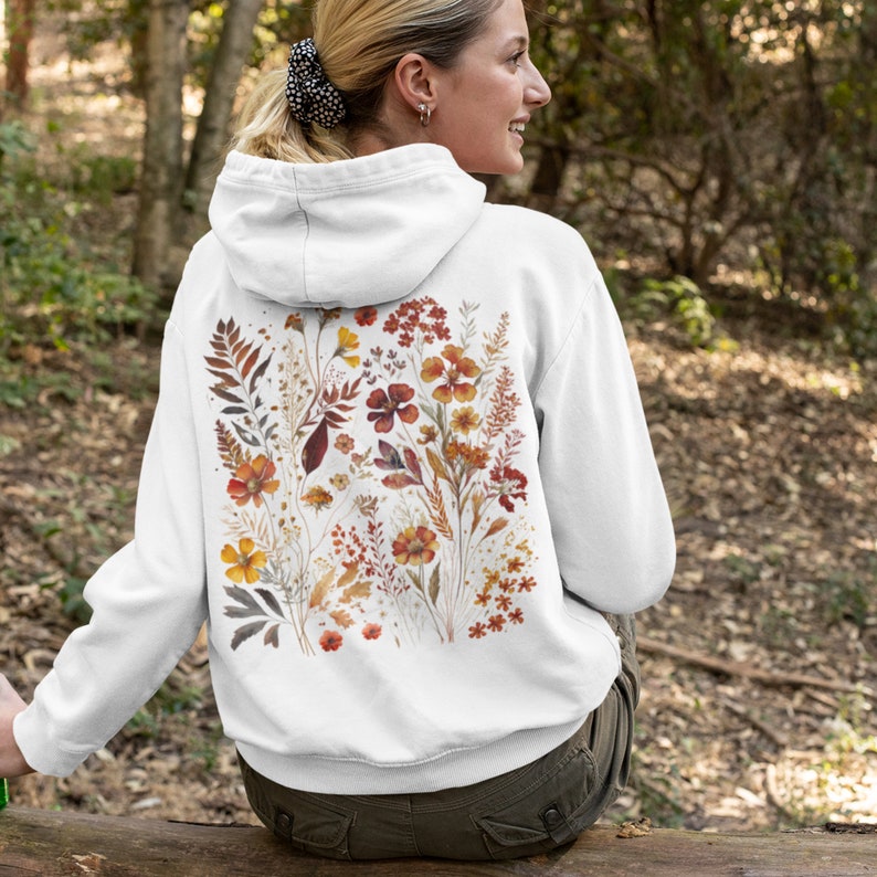 May include: White hoodie with a floral print on the back. The print features a variety of flowers and leaves in shades of orange, red, and yellow.