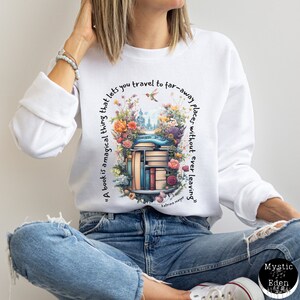 K&ouml;nnte beinhalten: Wei&szlig;es Sweatshirt mit einer bunten Grafik eines B&uuml;cherregals mit B&uuml;chern und Blumen. Der Text "Ein Buch ist eine magische Sache, die es dir erm&ouml;glicht, an ferne Orte zu reisen, ohne jemals zu gehen" ist auf dem Sweatshirt gedruckt.