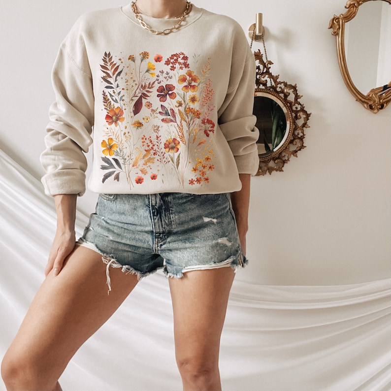 May include: A beige sweatshirt with a floral print featuring orange, yellow, and red flowers. The sweatshirt has long sleeves and a crew neck.