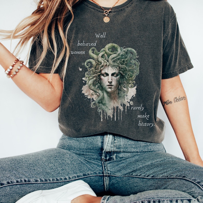 May include: Dark gray t-shirt featuring a Medusa graphic with green and white accents. The shirt has the text "Well behaved women rarely make history."