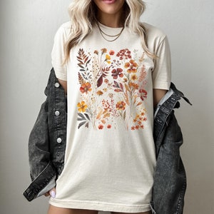 Puede incluir: Una camiseta beige con un estampado floral que presenta una variedad de flores en tonos naranja, rojo y amarillo. Las flores est&aacute;n dispuestas en un racimo en la parte delantera de la camiseta.
